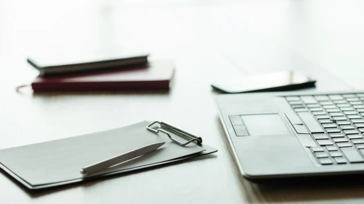 Very bright picture of a work desk with soft reflections and a blurry, almost vanishing, background.
                  Within the scene there's a business laptop, a smartphone, a pen, two notebooks and a clipboard.